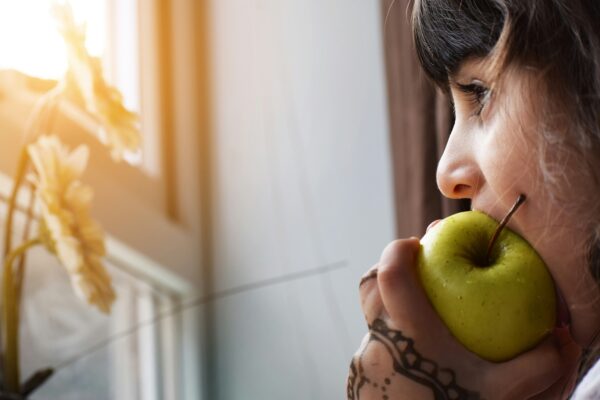 A close-up image of a young girl with henna tattoos on her hand eating a green apple looking out of a sunny window